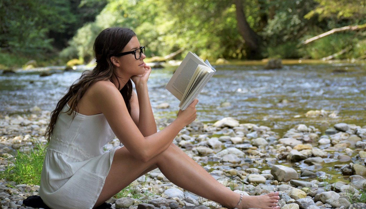 Woman in glasses reading book outside by river